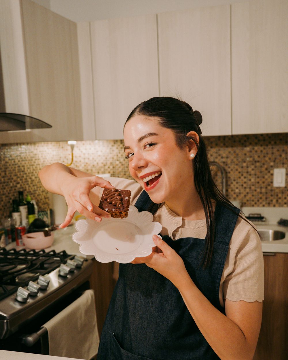 Marianna riendo mientras ofrece a la cámara un brownie servido en un plato blanco, con la tabla de brownies enfrente.