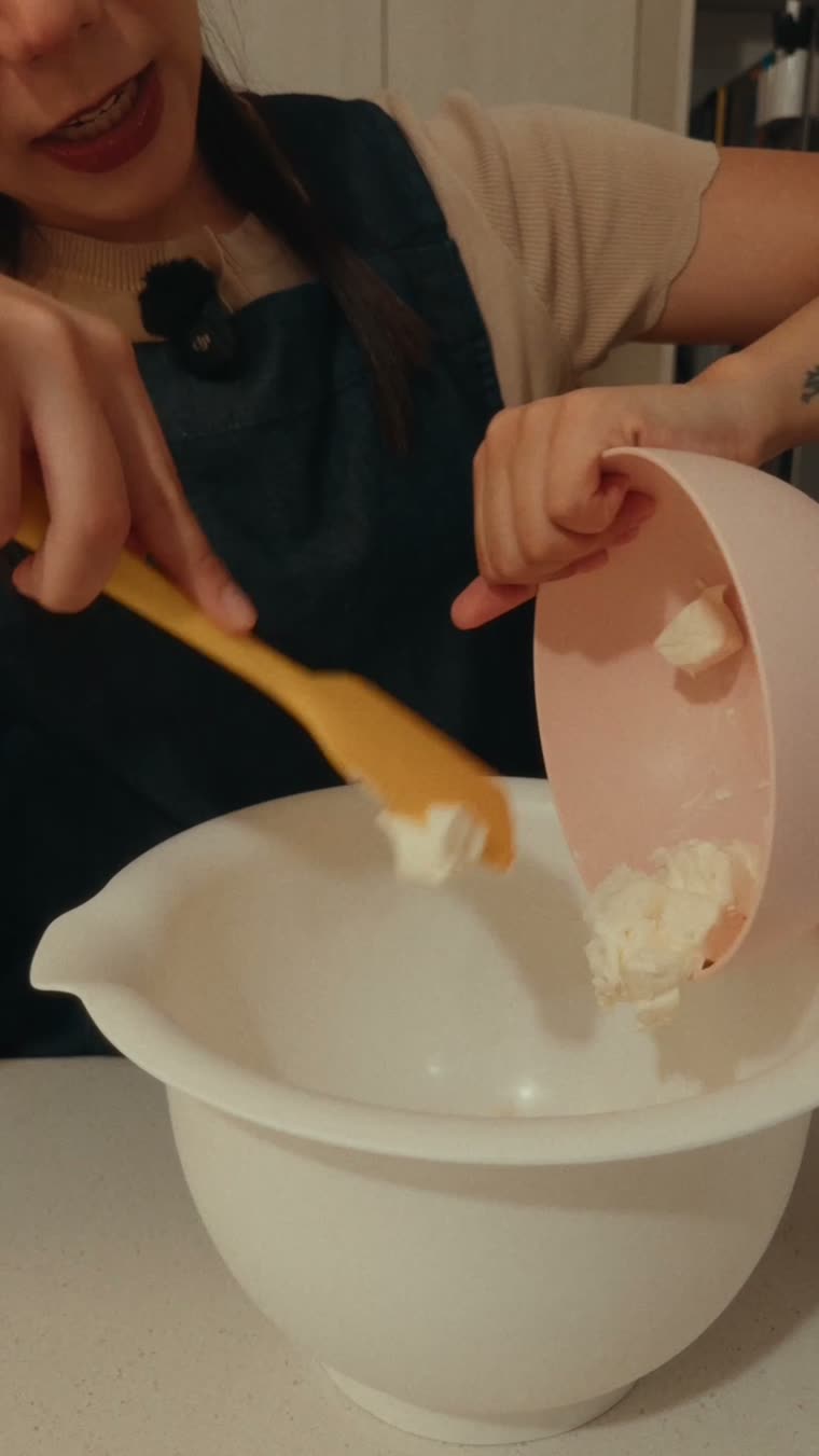 Marianna pasa el queso crema del tazón rosa al tazón grande con una espátula.